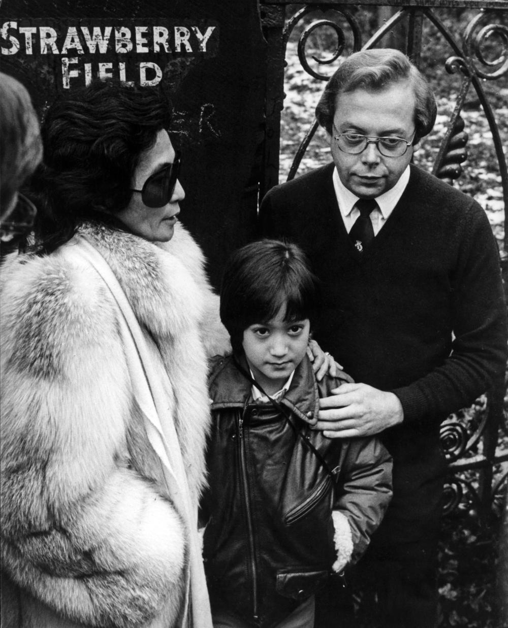 Yoko Ono and a young Sean Ono Lennon and a third man stand in front of a gate post which says Strawberry Field