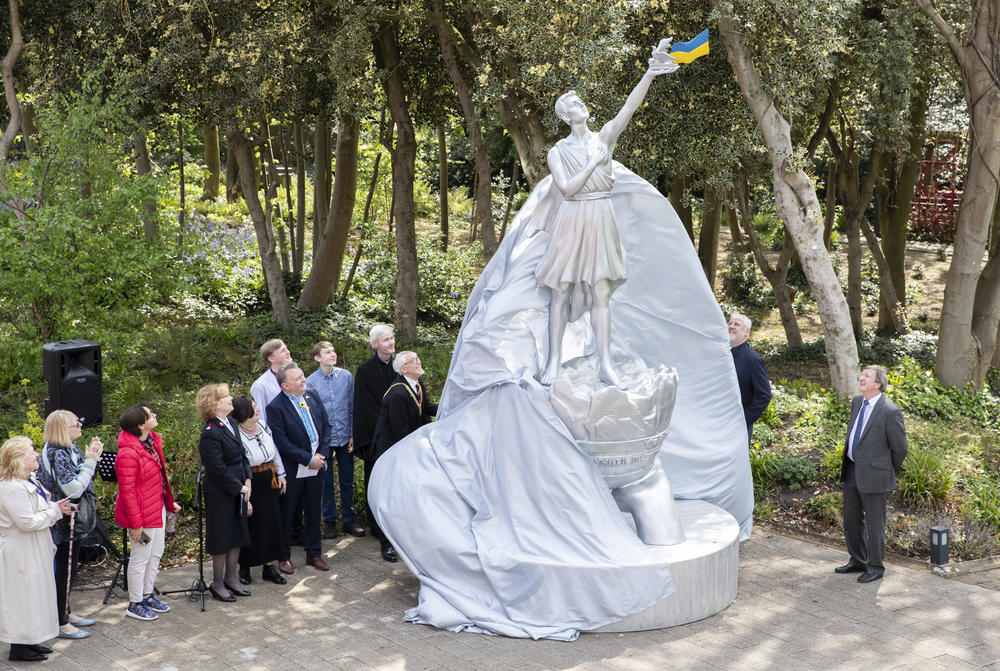 A large aluminium statue is unveiled by a group of people in a garden